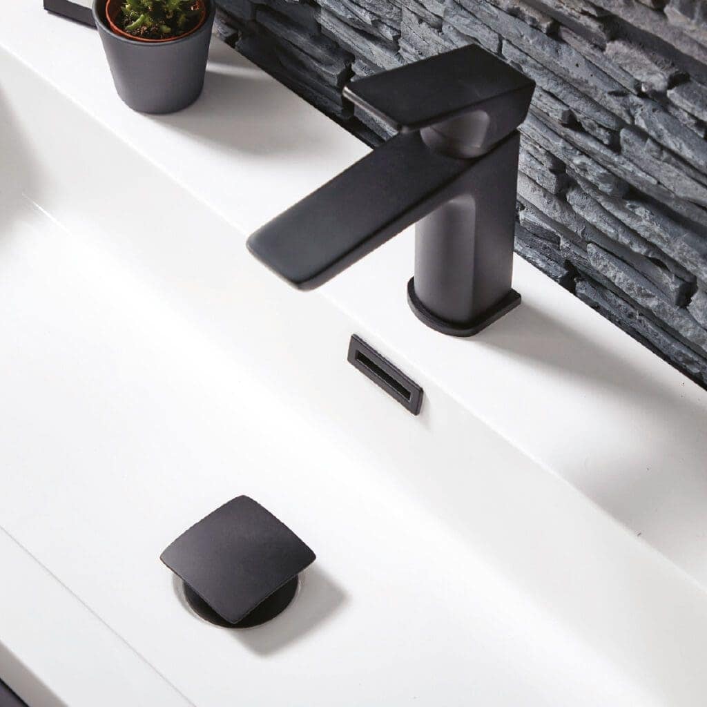 Sleek black bathroom tap with matte finish on a modern white sink, complemented by a matching drain cover, with textured stone wall and a small potted plant for stylish bathroom decor.