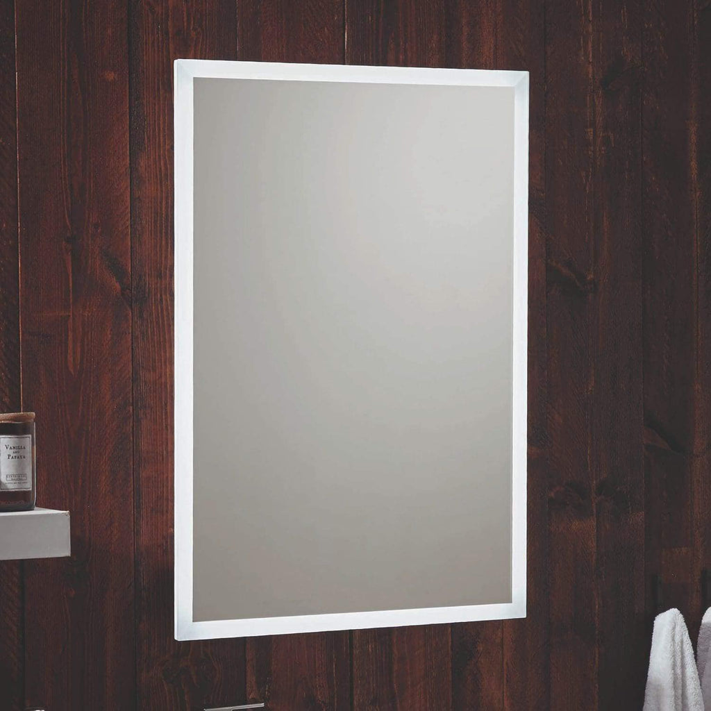 Clean bathroom mirror with white frame mounted on wooden wall, reflecting light, with a shelf holding a vanilla scented candle and a towel hanging below.