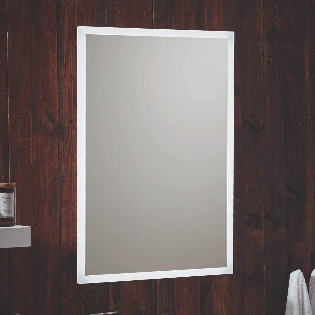 Clean bathroom mirror with white frame mounted on wooden wall, reflecting light, with a shelf holding a vanilla scented candle and a towel hanging below.