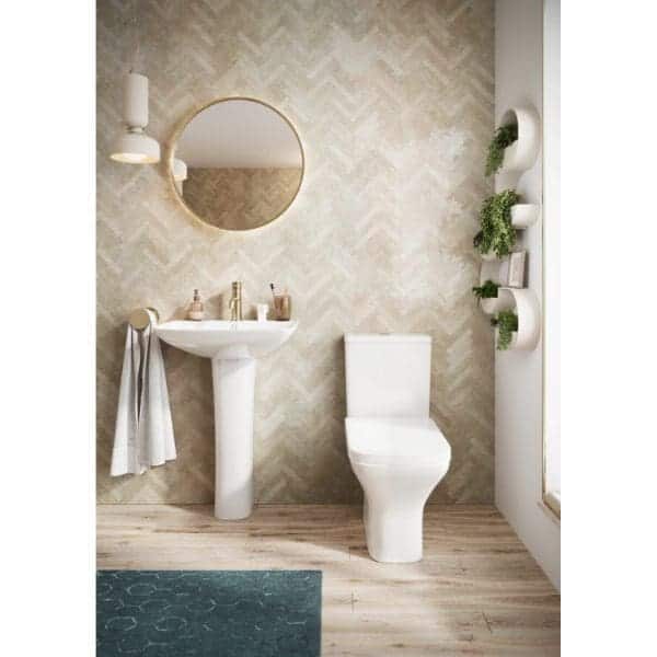 Minimalist bathroom with pedestal sink, round mirror, and modern toilet, featuring stylish wall-mounted lighting and green foliage decor accents, creating a fresh and contemporary bathroom design.