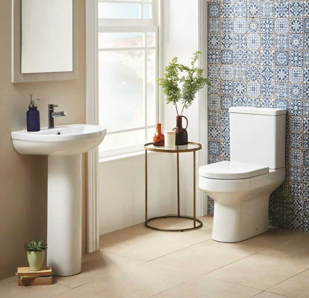 Modern bathroom with white ceramic toilet and pedestal sink, decorative blue patterned tiles, and natural light from large window, creating a stylish and functional space.