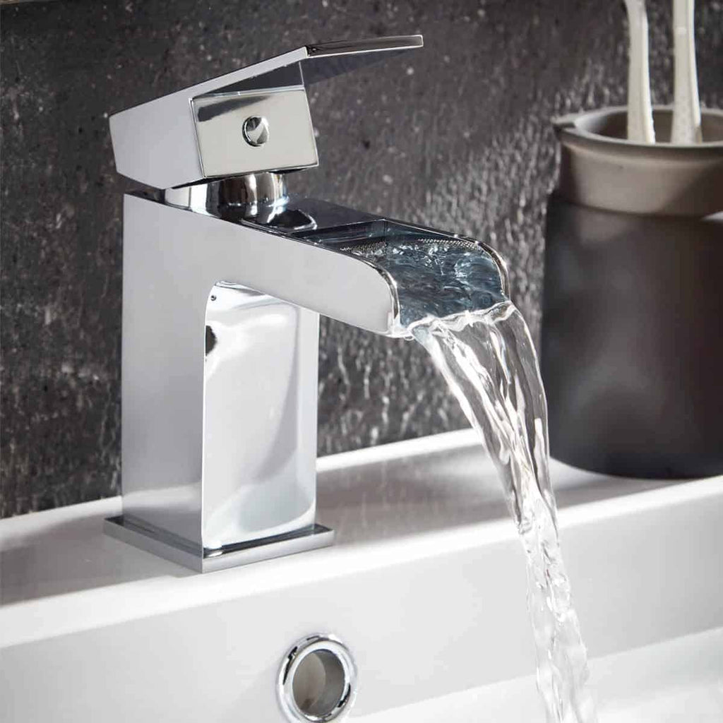 Sleek modern chrome bathroom tap with waterfall feature flowing into a white ceramic sink on a textured grey wall background.