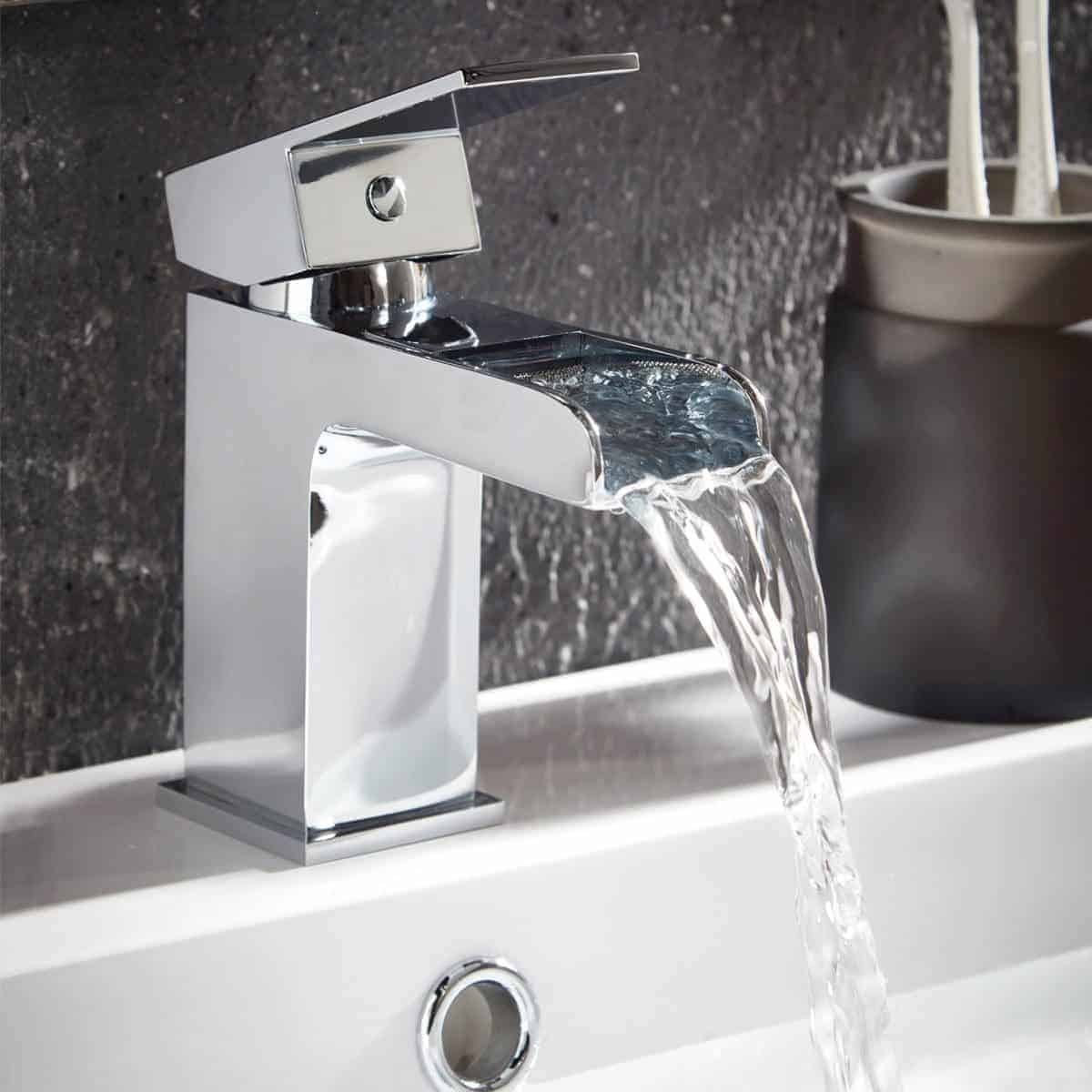 Sleek modern chrome bathroom tap with waterfall feature flowing into a white ceramic sink on a textured grey wall background.